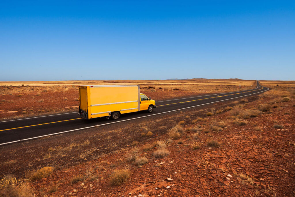 A yellow moving truck driving on an empty desert road.