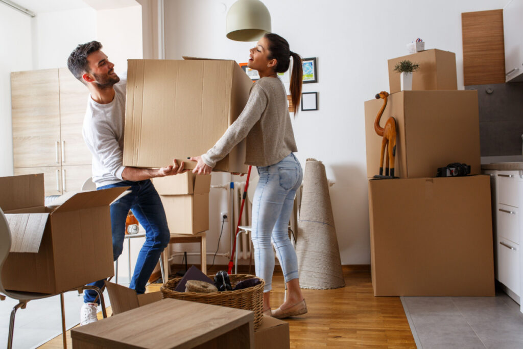 A couple lifting a cardboard box; there are other cardboard boxes stacked throughout the room.